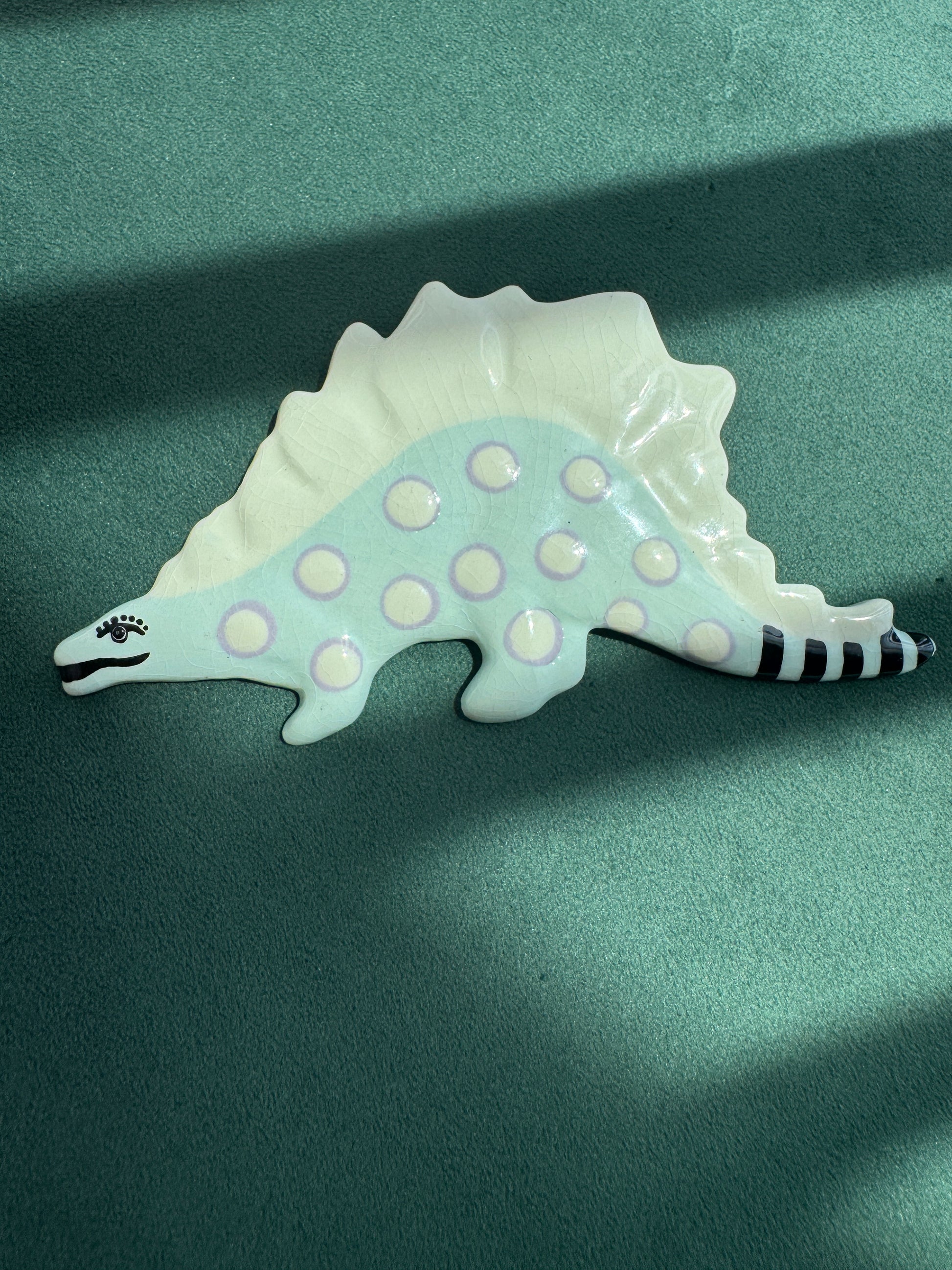 A ceramic Stegosaurus dinosaur figurine with a pale mint green body, white spikes and feet, and black-and-white striped tail details, displayed on a textured dark green surface.