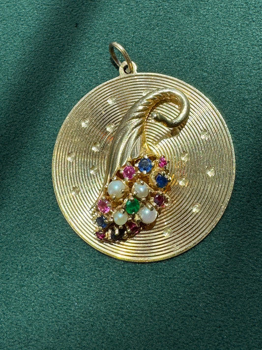 A round gold pendant featuring a textured concentric circle design, a curved gold leaf motif, and a central cluster of multicolored gemstones including white pearls, pink, blue, green, and red stones, displayed on a dark green velvet background.