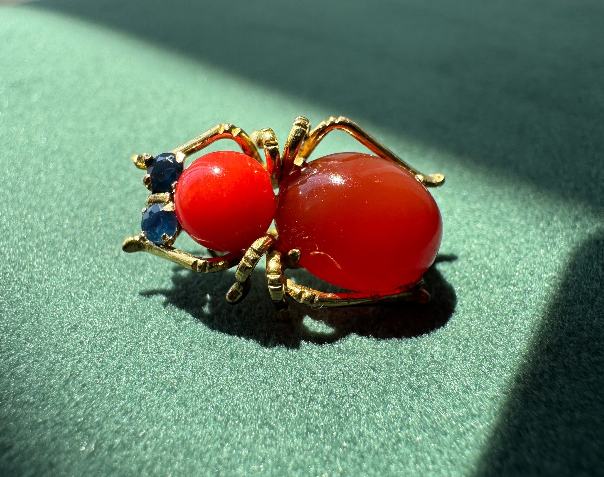 A gold spider brooch with a large red cabochon body, a smaller red eye, and two small blue gemstone accents on its legs, resting on a green textured surface.