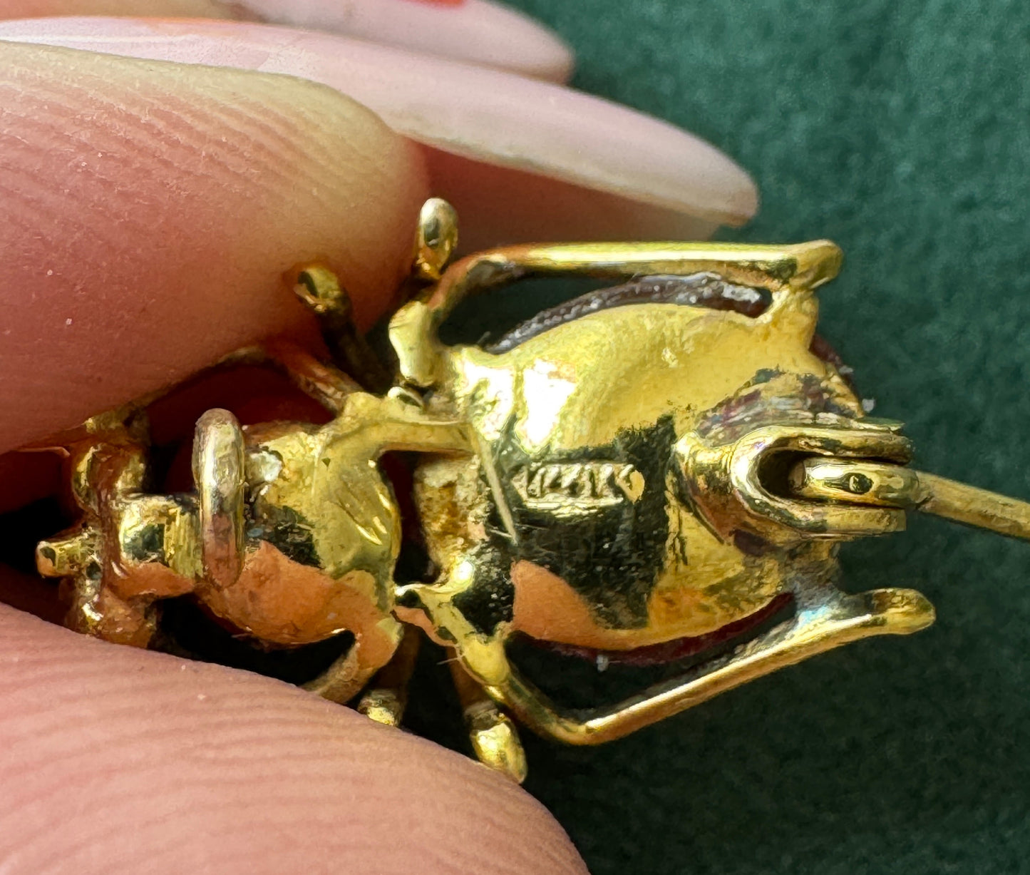 A close-up of a gold beetle-shaped brooch held between fingers against a dark green textured background.