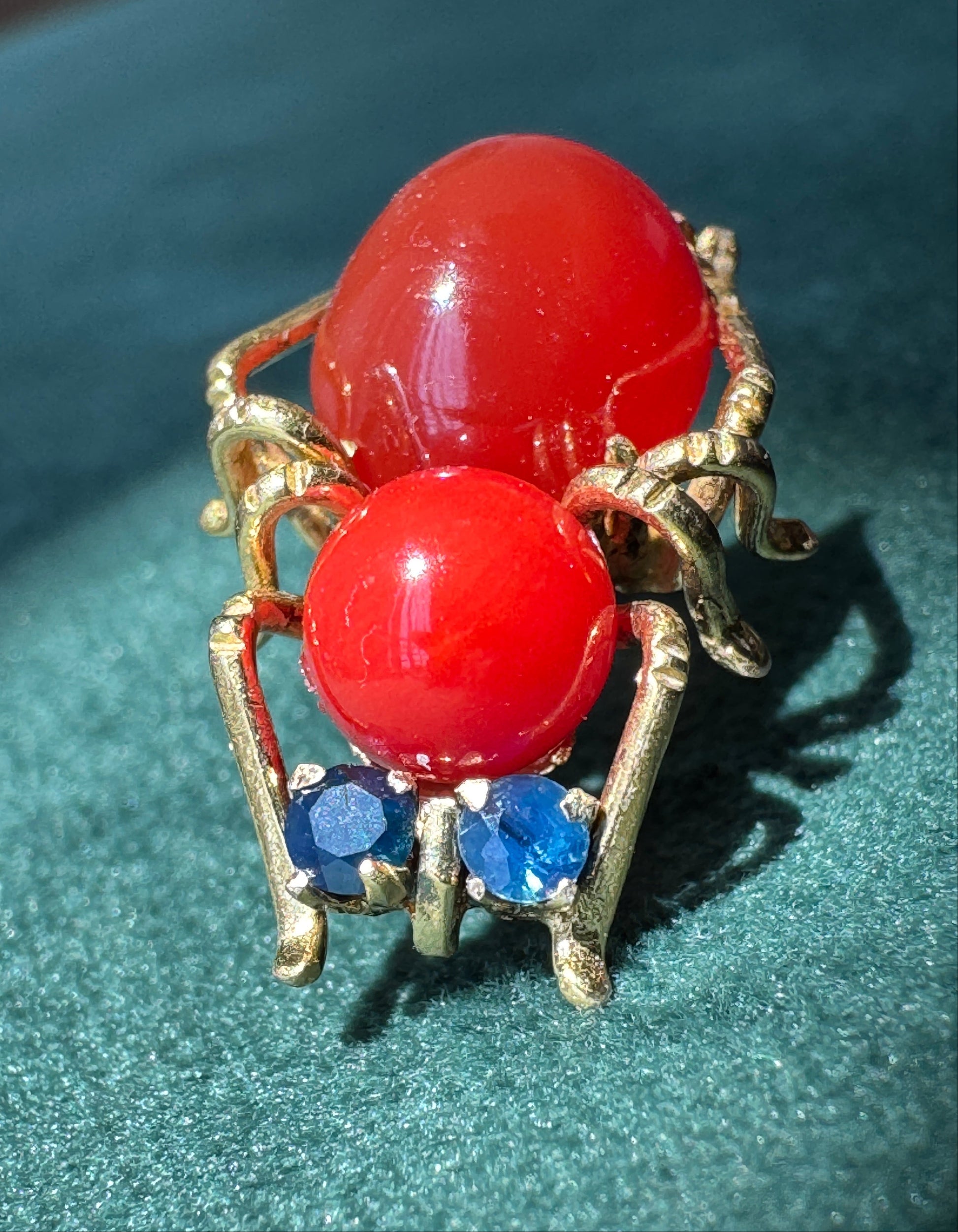 A spider-shaped ring featuring two large glossy red oval stones for eyes, three small blue round stones for legs, and gold-toned metal for the body and legs, displayed on a textured dark green surface.