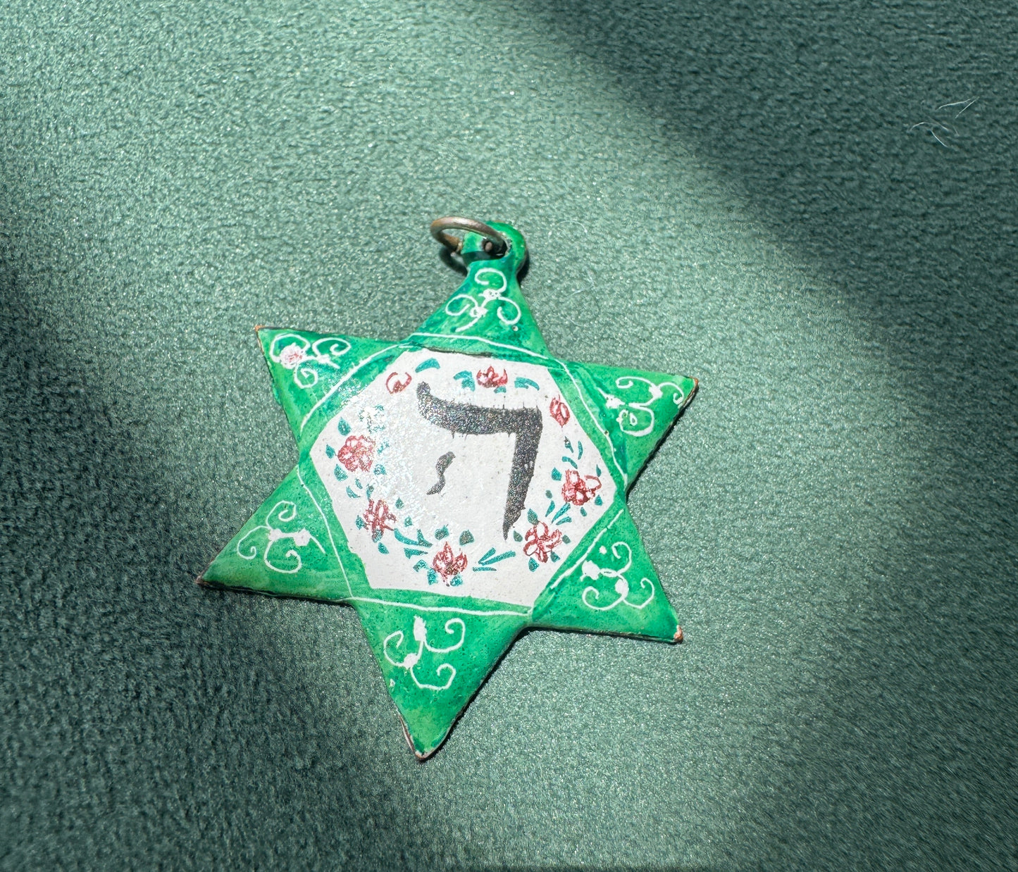 A handmade green paper Star of David pendant with a white center, floral details, and a black Hebrew letter, resting on a textured green surface with diagonal shadows.