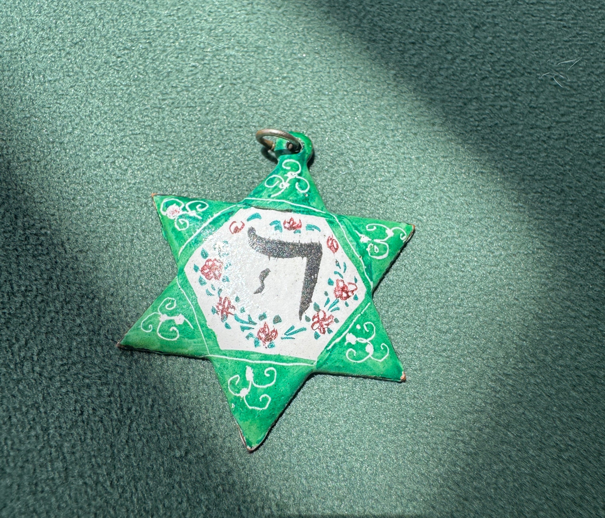 A handmade green paper Star of David pendant with a white center, floral details, and a black Hebrew letter, resting on a textured green surface with diagonal shadows.