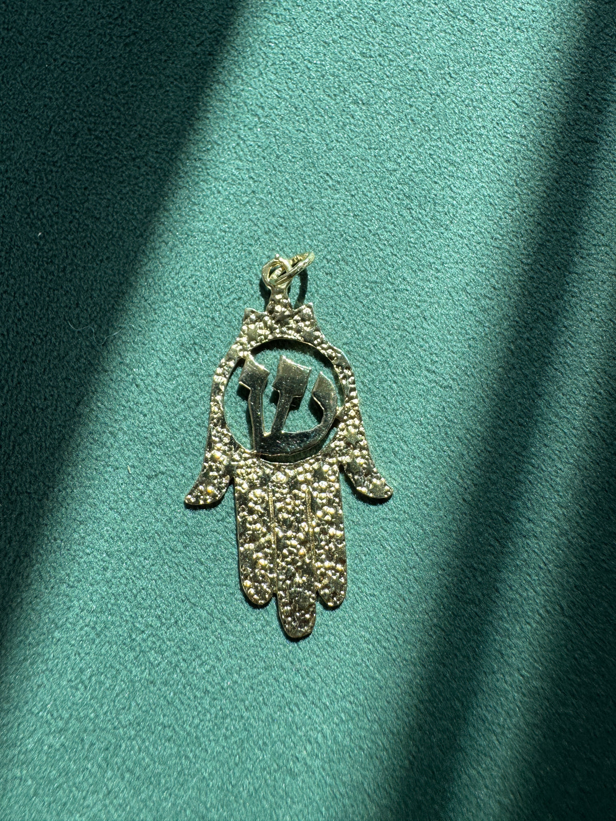 A textured gold hamsa hand pendant with a central circular cutout, resting on a teal-green textured surface with diagonal light and shadow patterns.